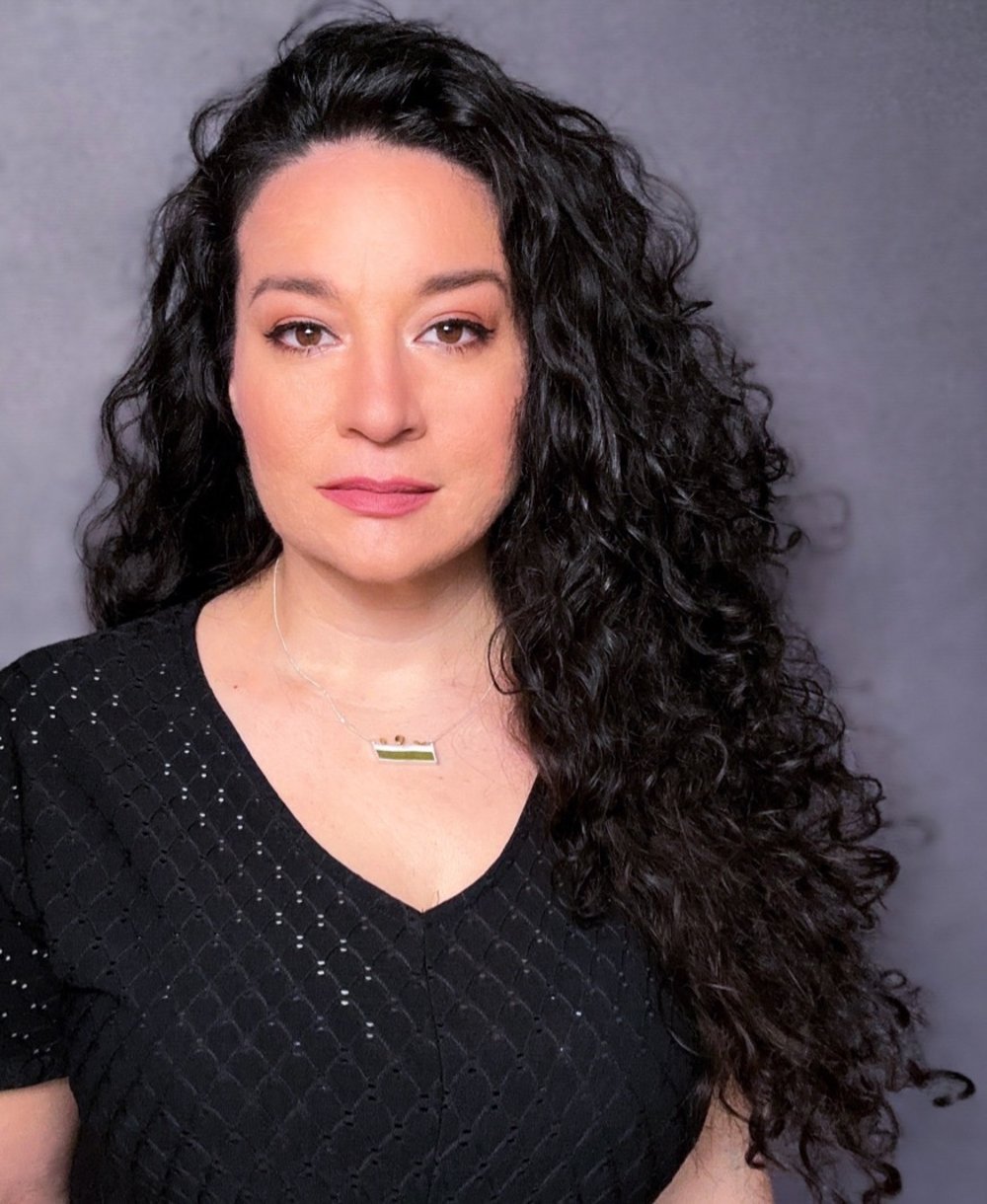 a woman with dark curly hair wearing a black shirt looks at the camera